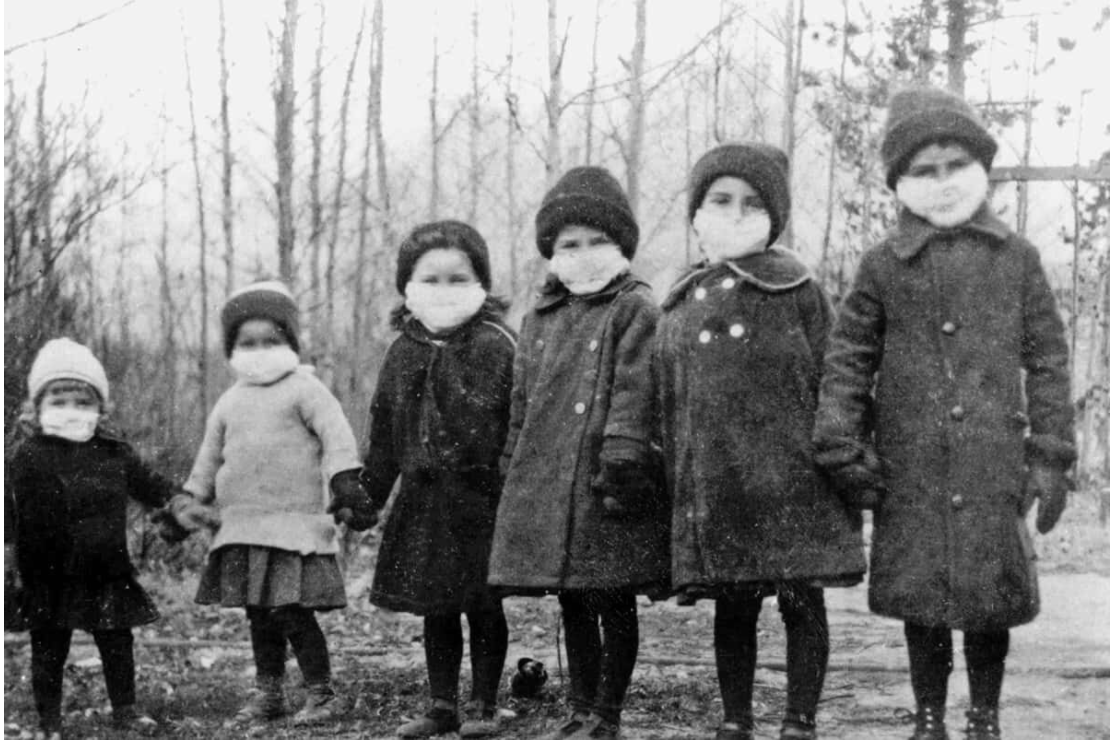 An archival image of of six children standing in winter clothing wearing face masks.