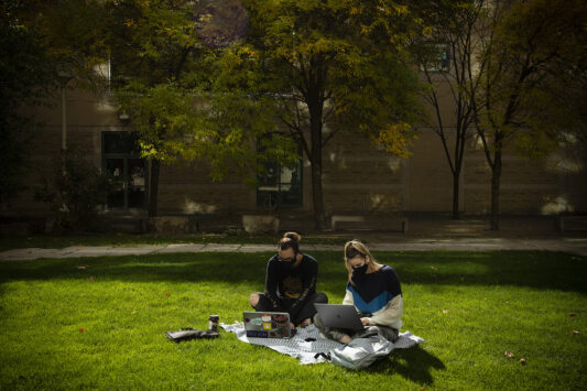 Two students outside in masks working on their laptops on a picnic blanket.