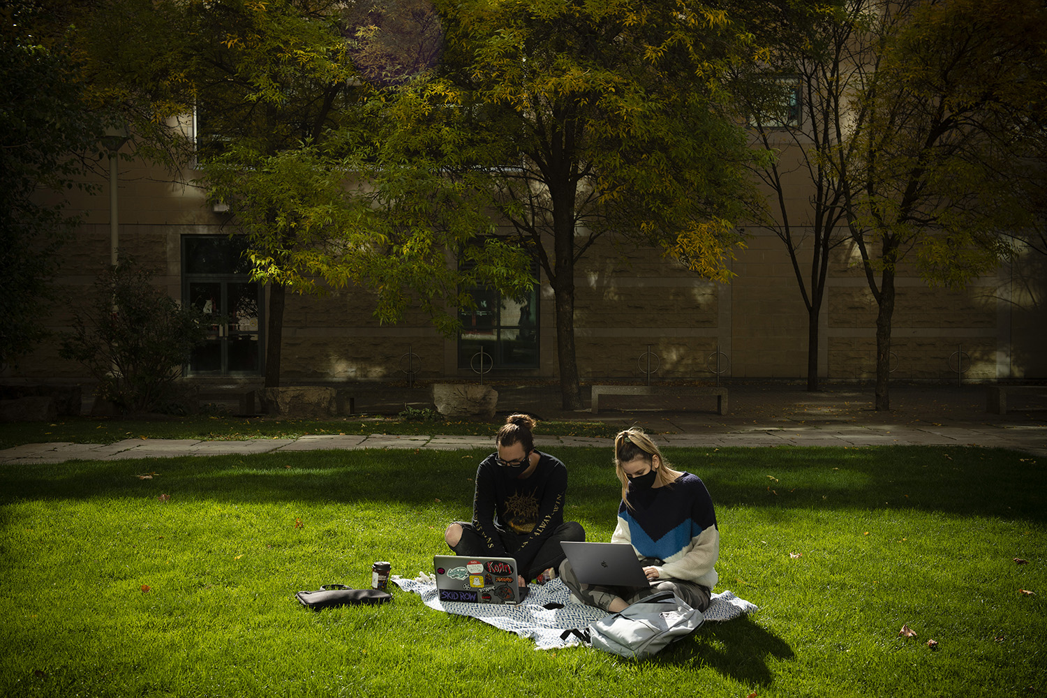Two students outside in masks working on their laptops on a picnic blanket.