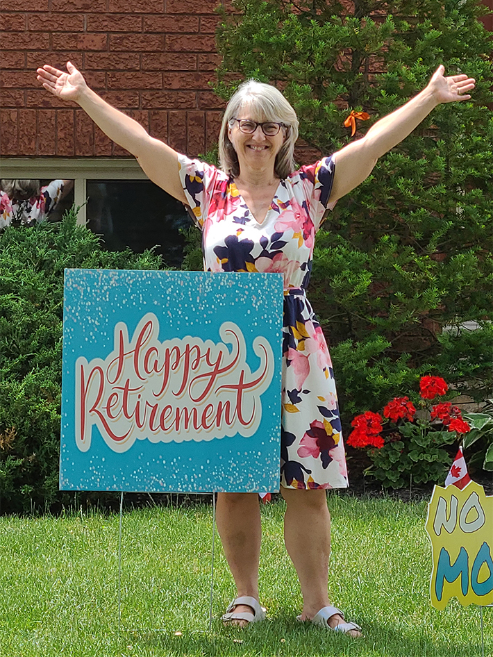 A woman stands outside with a sign saying "happy retirement" on her lawn.