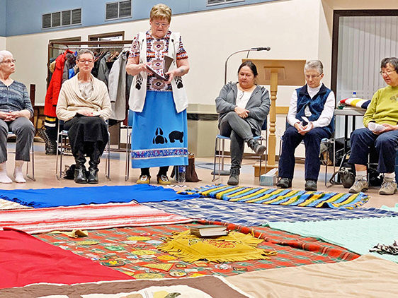 Four people sit in chairs around blankets placed on the floor, one person stands, reading from a book.