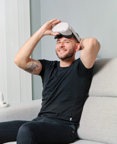 Young man in black t-shirt and jeans seated on a grey couch and lifts a pair of VR goggles up to his forehead as he smiles at the camera.