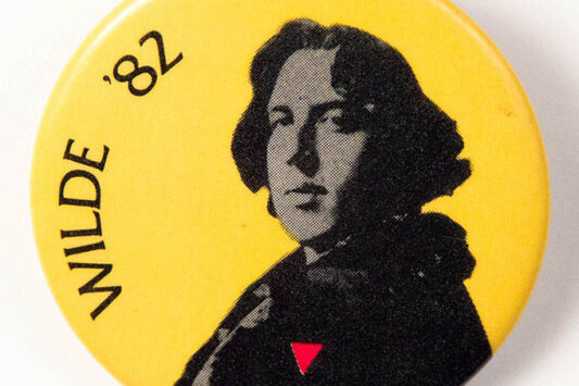 Circular yellow button with a black and white image of Oscar Wilde wearing an upside down pink triangle instead of his usual carnation.