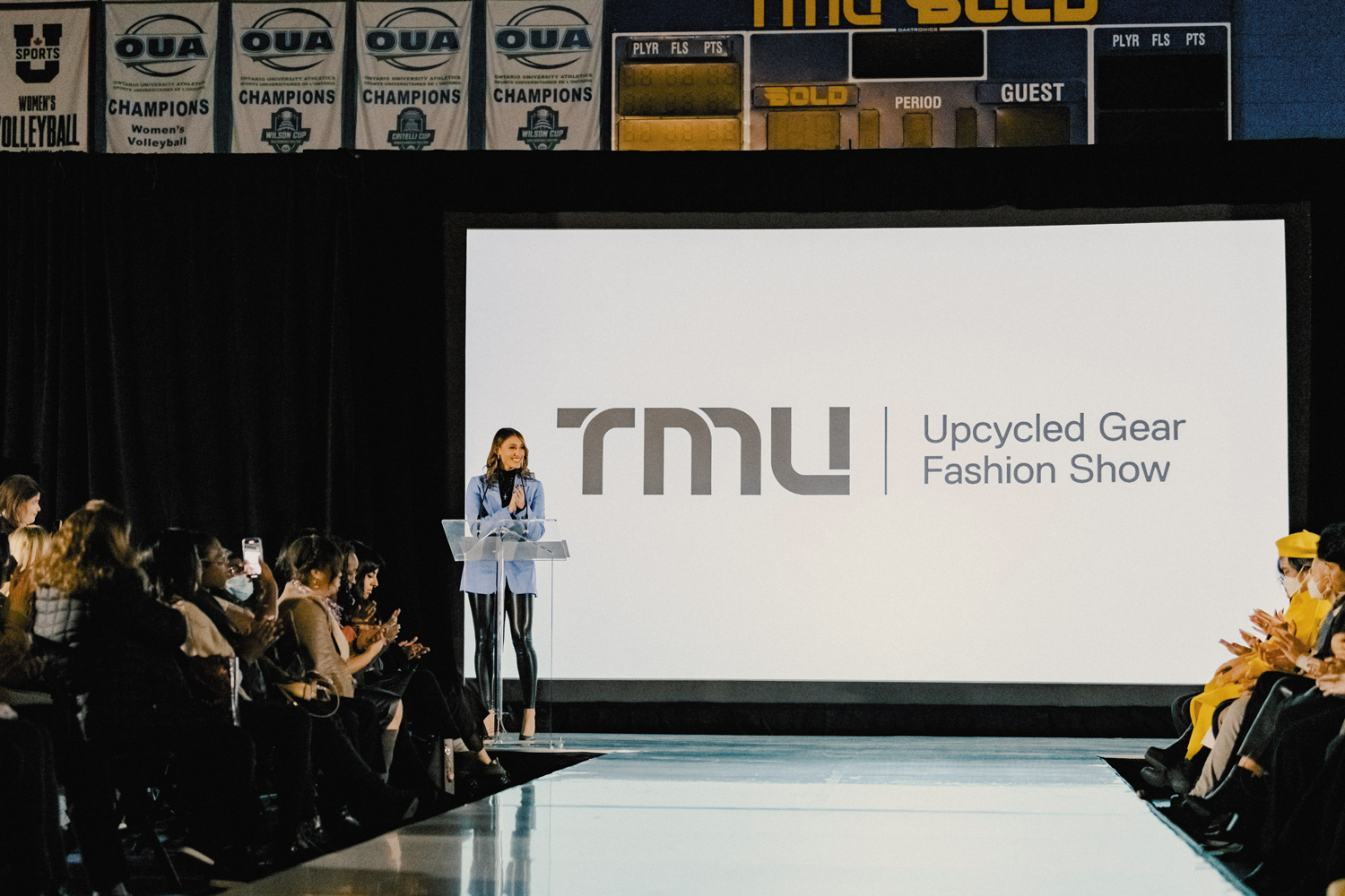 A woman stands at a podium in front of a large screen that says TMU Upcycled Gear Fashion Show
