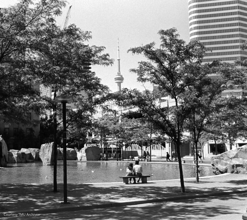 A couple sits on a bench looking at Lake Devo, with the CN Tower in the distance.