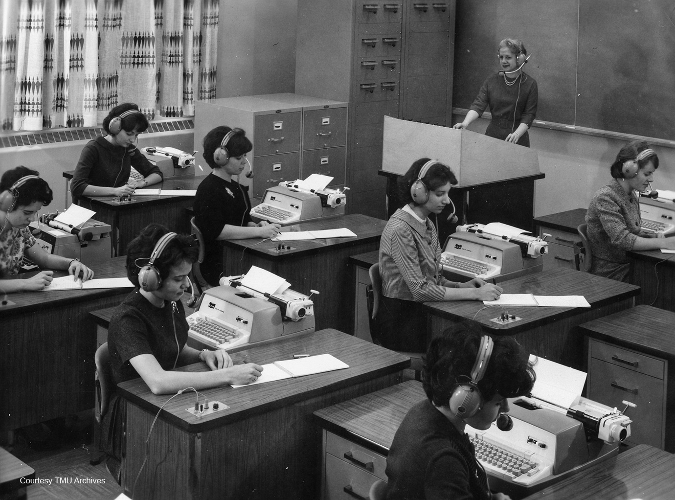 Women wearing headphones sitting at desks with typewriters.