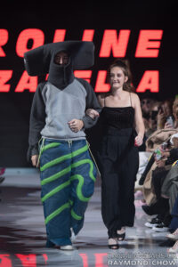 Karoline Mazzarella walking down the runway in an all black ensemble with a model dressed in Karoline’s design.