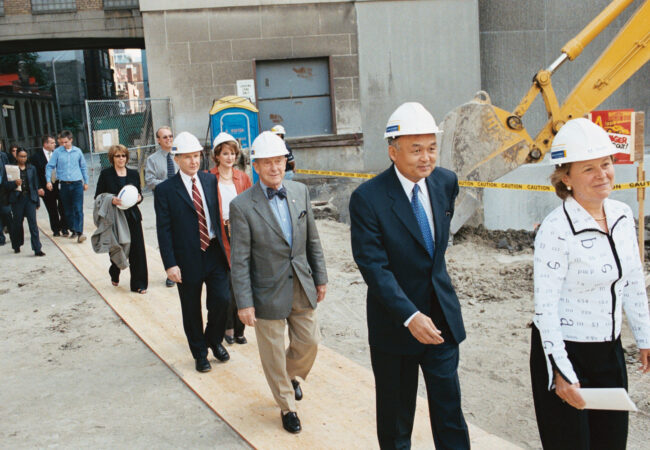 University administrators, including G. Raymond Chang, wearing hardhats walk single file to the Heaslip House building groundbreaking. A backhoe operates in the background.