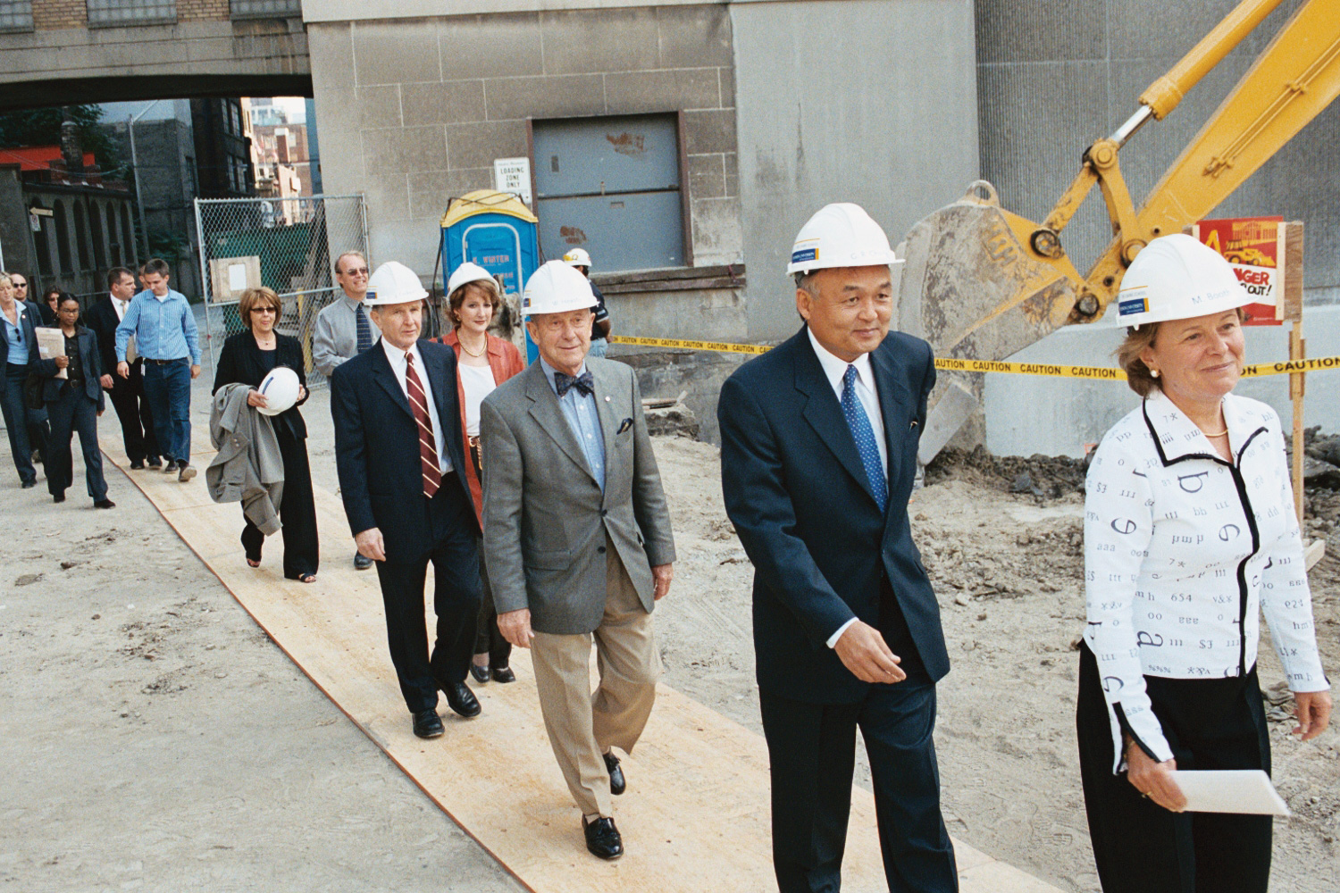 University administrators, including G. Raymond Chang, wearing hardhats walk single file to the Heaslip House building groundbreaking. A backhoe operates in the background.