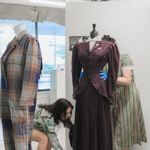 Two researchers dressing a mannequin in a two-piece checked purple wool womenswear suit. One student stands behind it with their hands on the waist, the other bends to touch the bottom of the suit.