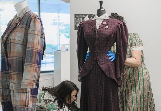 Two researchers dressing a mannequin in a two-piece checked purple wool womenswear suit. One student stands behind it with their hands on the waist, the other bends to touch the bottom of the suit.