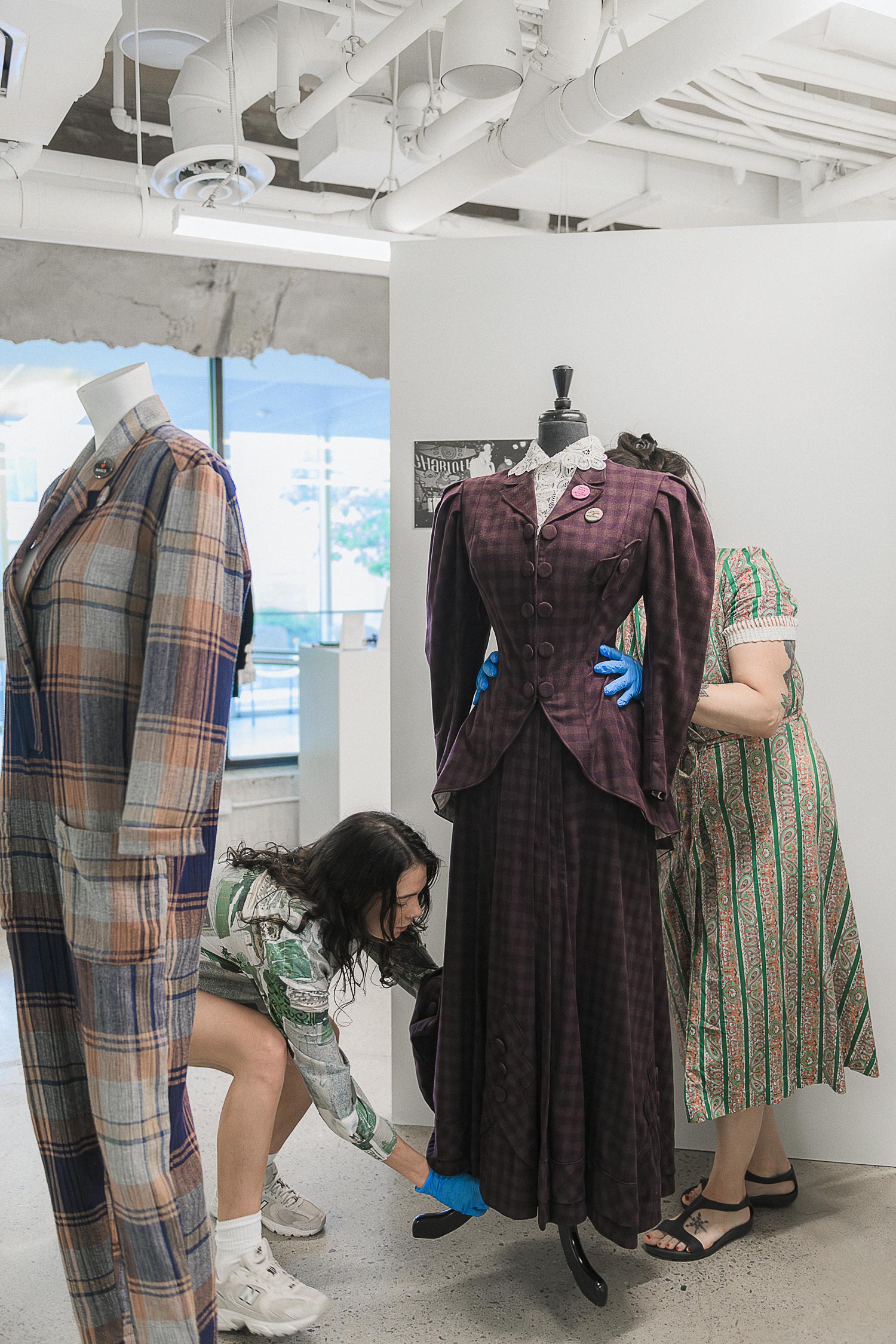 Two researchers dressing a mannequin in a two-piece checked purple wool womenswear suit. One student stands behind it with their hands on the waist, the other bends to touch the bottom of the suit.