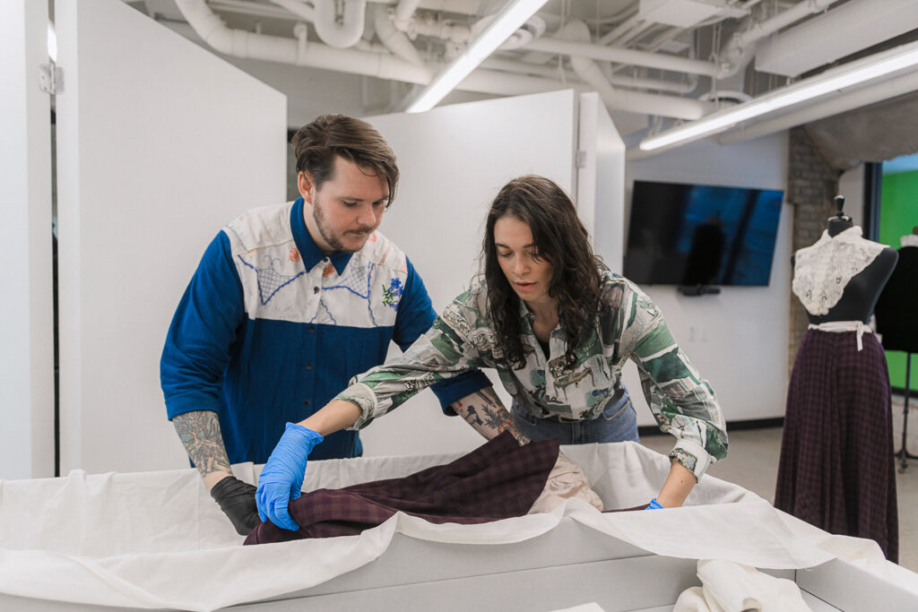 Patrick and Sephra carefully lay a dress into a garment box. They are wearing protective gloves.