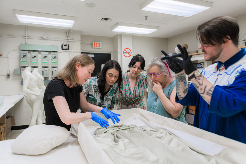 Eve Townsend, the director of the FRC delicately touches a dress in a garment box, she is wearing blue gloves. Four other researchers from the FRC watch.