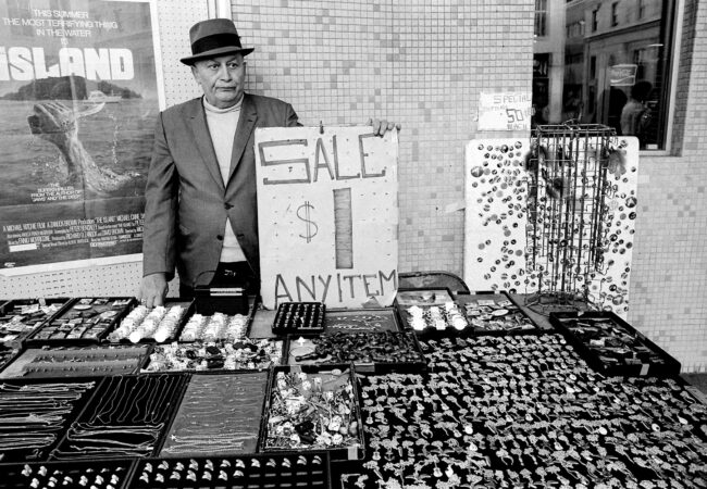 A man wearing a hat stands behind a table of jewelry for sale. A sign says $1 any item.