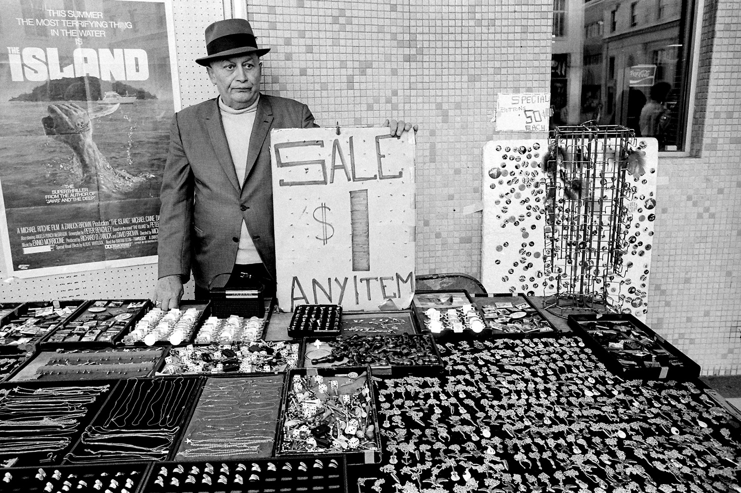 A man wearing a hat stands behind a table of jewelry for sale. A sign says $1 any item.