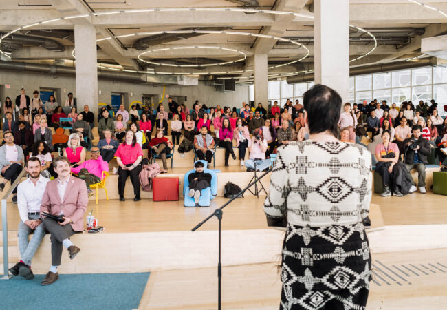 Chelazon LeRoux, Two-Spirit artist and Canada's Drag Race contestant, at a podium speaking to a large crowd in pink shirts.