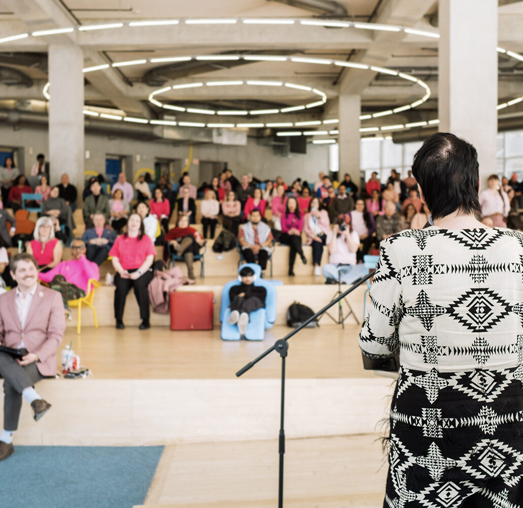 Chelazon LeRoux, Two-Spirit artist and Canada's Drag Race contestant, at a podium speaking to a large crowd in pink shirts.