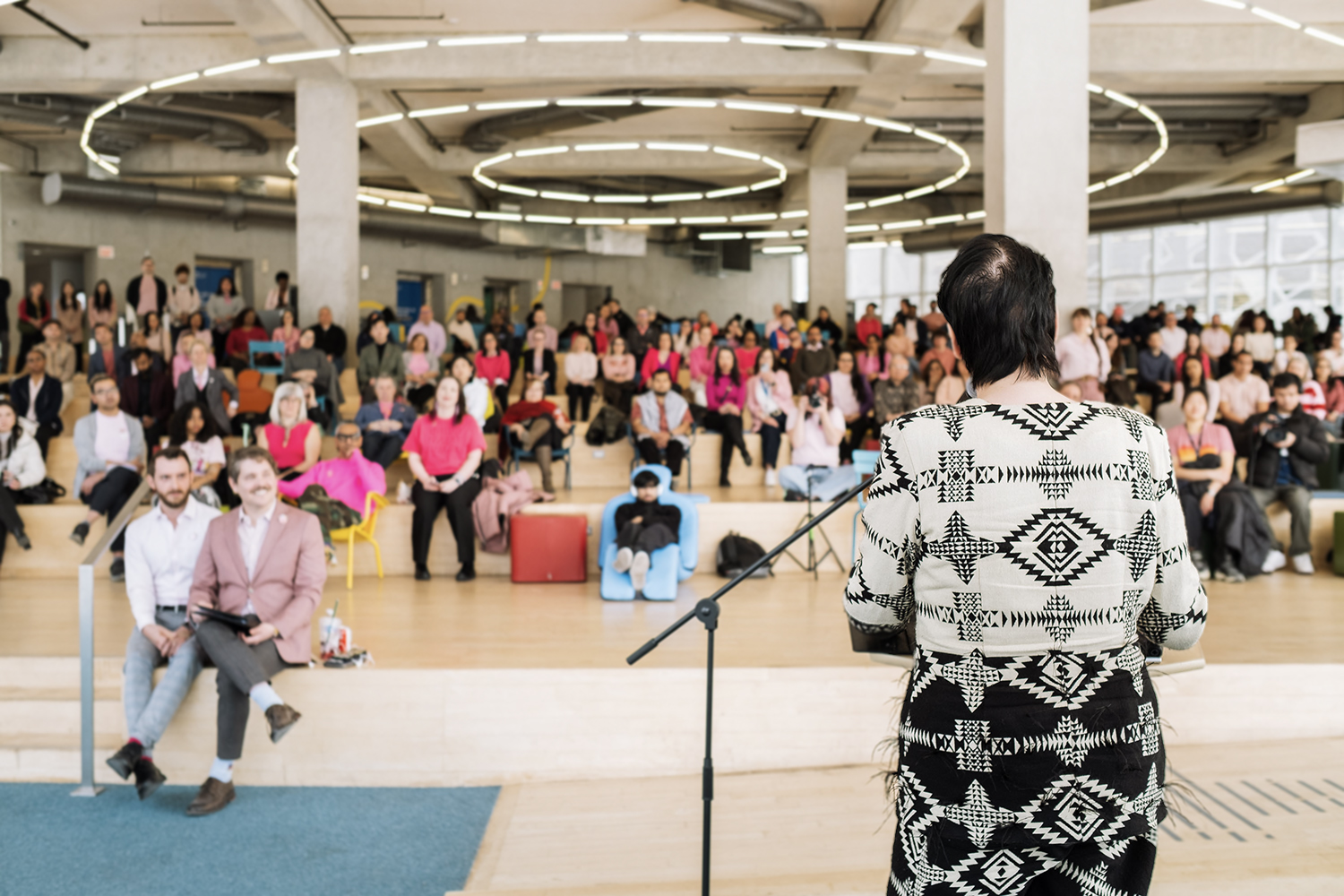 Chelazon LeRoux, Two-Spirit artist and Canada's Drag Race contestant, at a podium speaking to a large crowd in pink shirts.