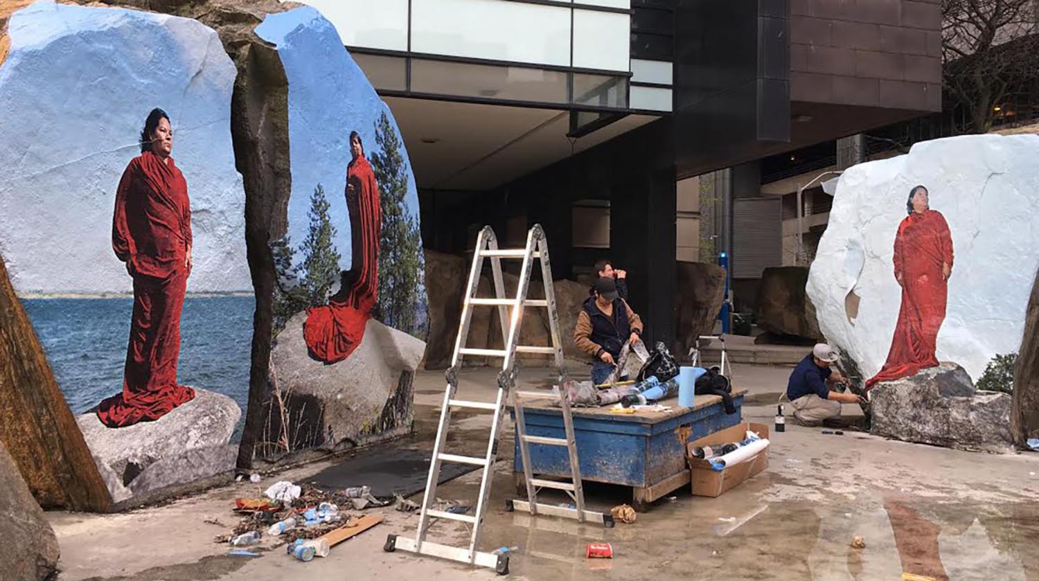 Installation of photographic images on the boulders in Lake Devo depicting the artist draped in a red robe, standing atop a boulder, in front of a lake.