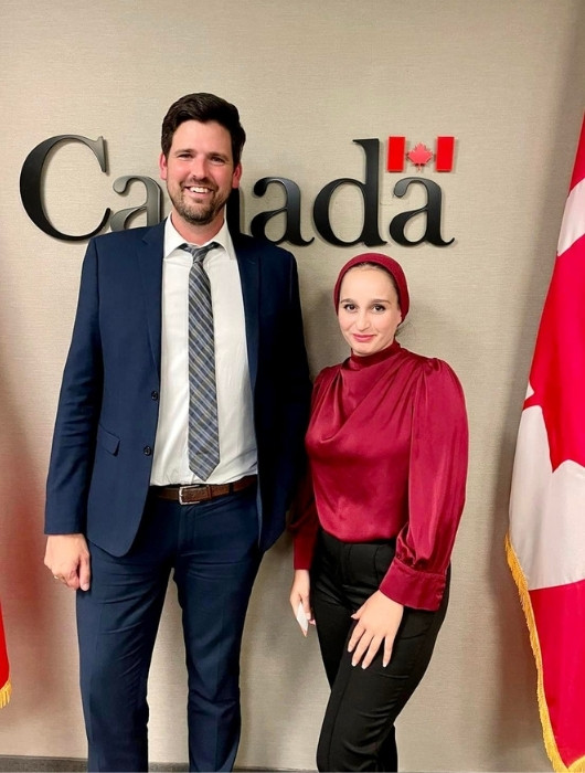 Sean Fraser, wearing a suit, stands next to TMU grad Hanen Namaa, with a Canadian flag to the right of the duo.
