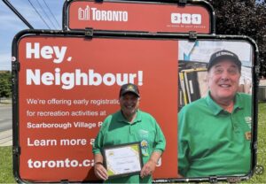 A man in a green t-shirt and black hat that says City of Toronto stands in front of an orange sign.
