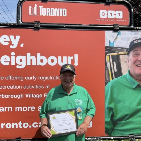 A man in a green t-shirt and black hat that says City of Toronto stands in front of an orange sign.