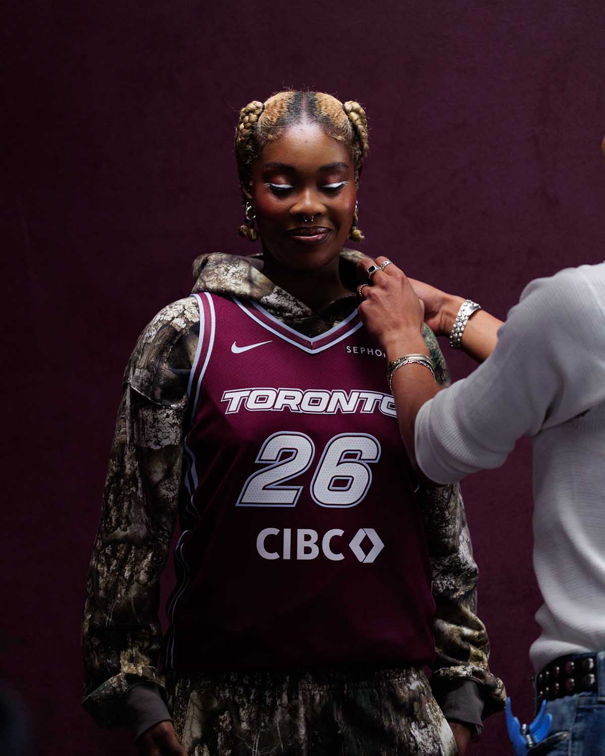 A person wearing a maroon Toronto Tempo jersey is styled on set, wearing a camo hoodie underneath.