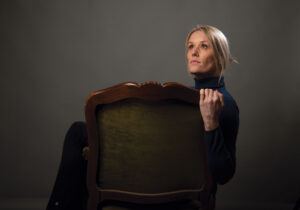 Dena Jackson poses in a studio portrait, holding the back of a chair in front of a dark neutral background.