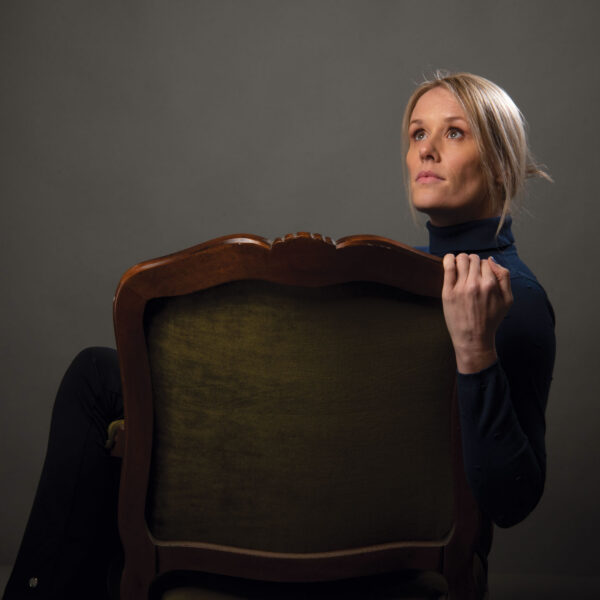 Dena Jackson poses in a studio portrait, holding the back of a chair in front of a dark neutral background.