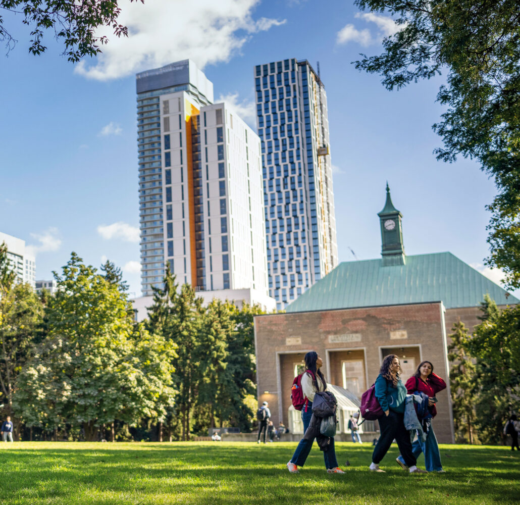 Students walk across a sunny campus green, with a historic clock-tower building and modern high-rises behind them.