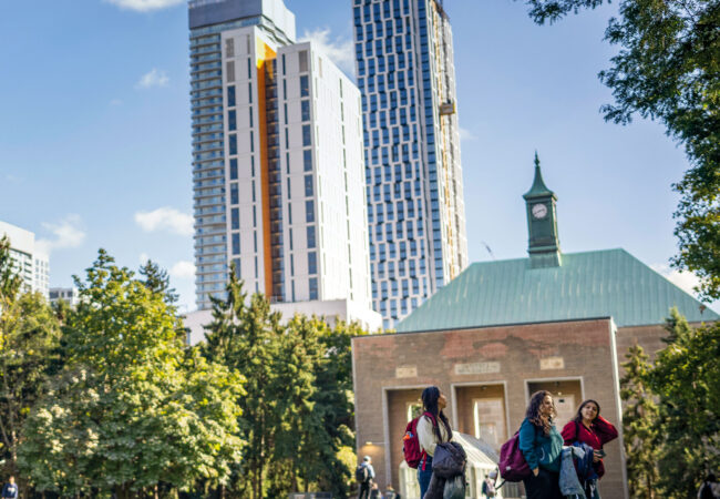 Students walk across a sunny campus green, with a historic clock-tower building and modern high-rises behind them.