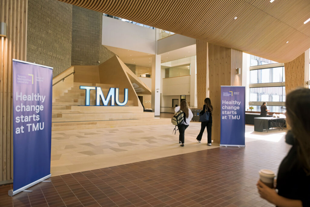 A bright TMU lobby with large illuminated letters on a staircase and banners reading "Healthy change starts at TMU."