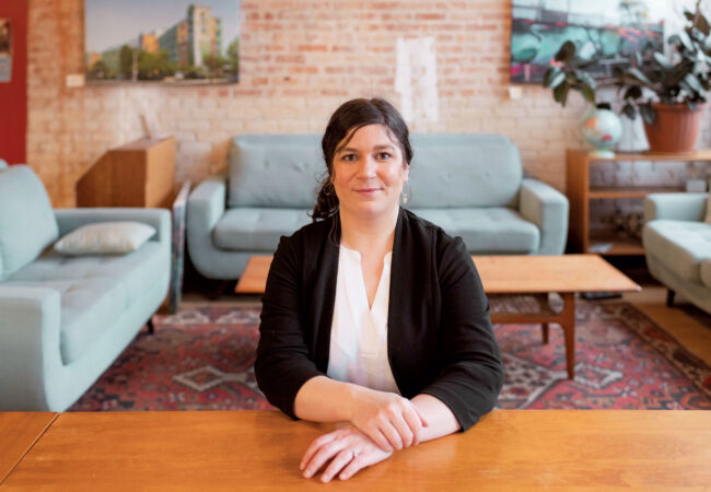 Desneige Frandsen seated at a wooden table in front of a couch, wearing a black cardigan and white blouse.