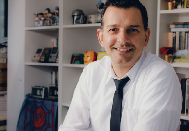 Sean Kheraj seated in an office, wearing a white shirt and tie and holding a green object.