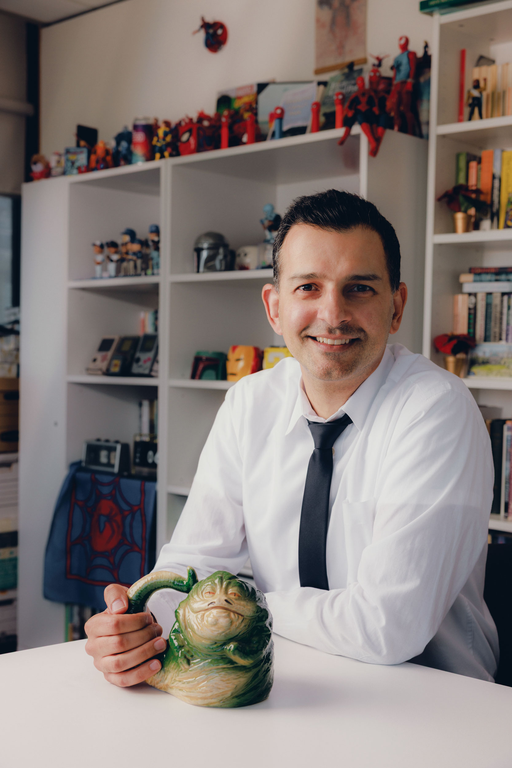 Sean Kheraj seated in an office, wearing a white shirt and tie and holding a green object.