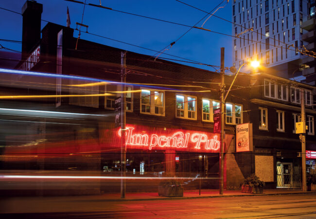 The Imperial Pub at night with its red neon sign glowing as a streetcar passes in front and the Library bar sign hangs on the building.
