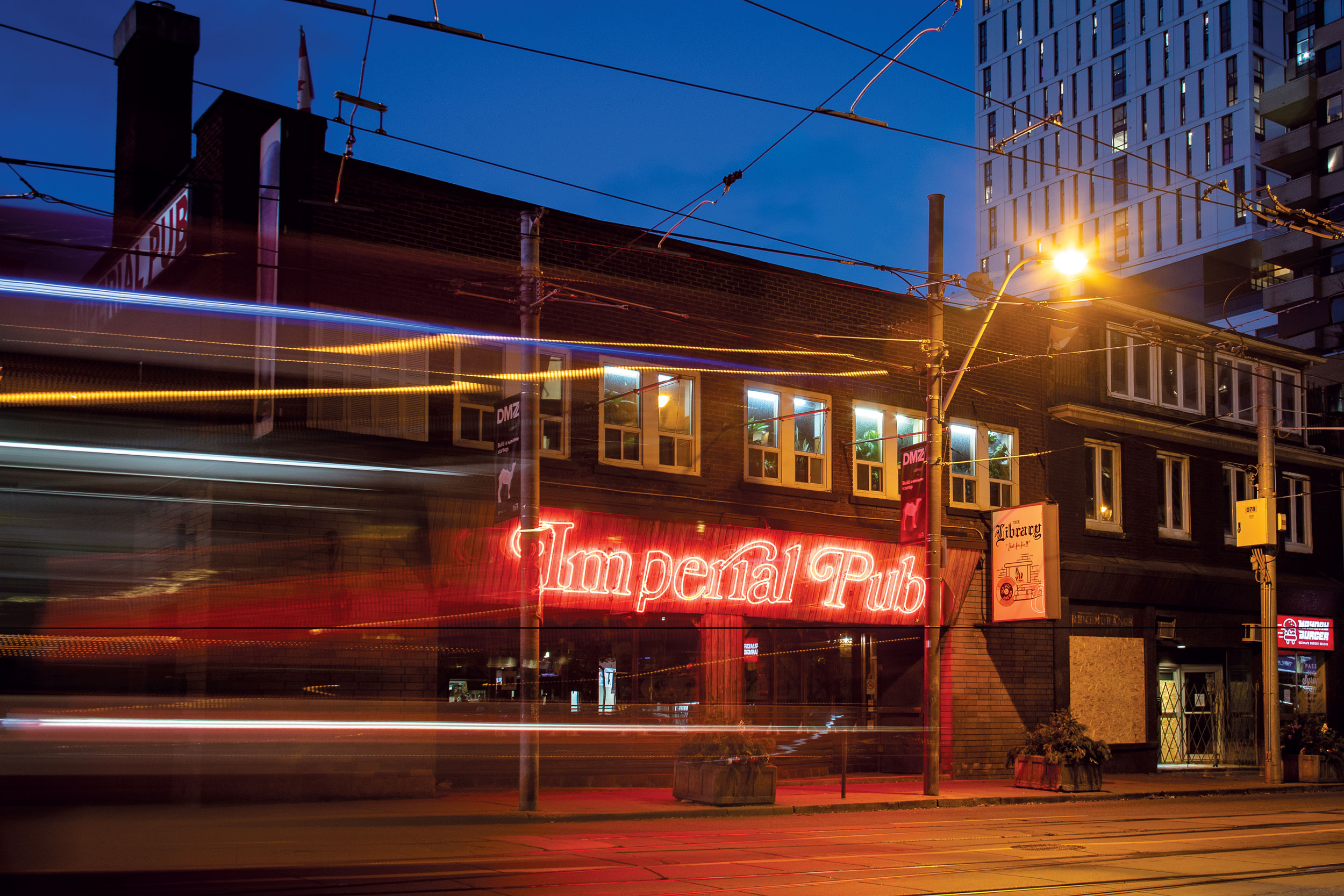 The Imperial Pub at night with its red neon sign glowing as a streetcar passes in front and the Library bar sign hangs on the building.
